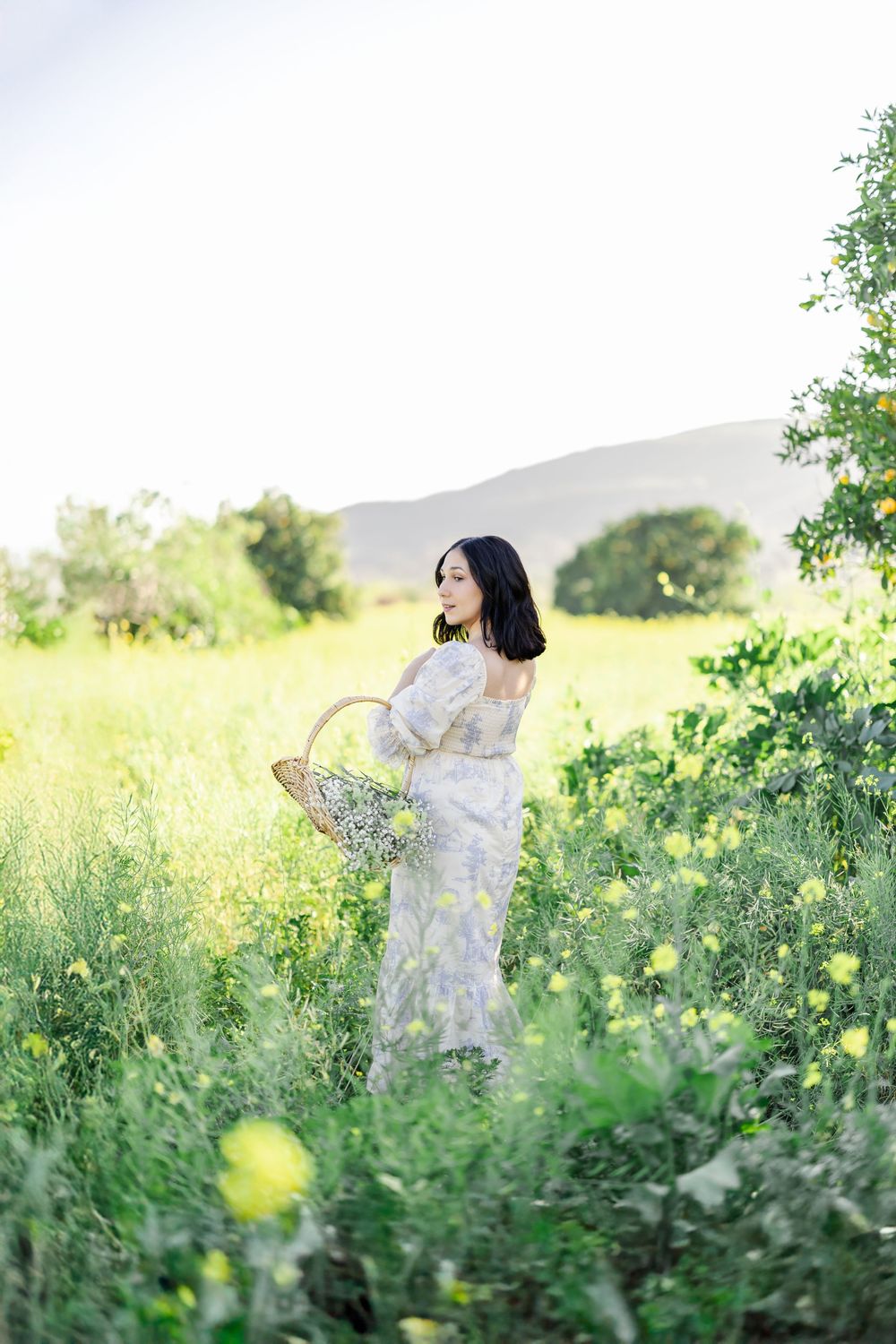 Woman in floral dress holding wicker basket stands in sunny green meadow with yellow wildflowers and mountains.