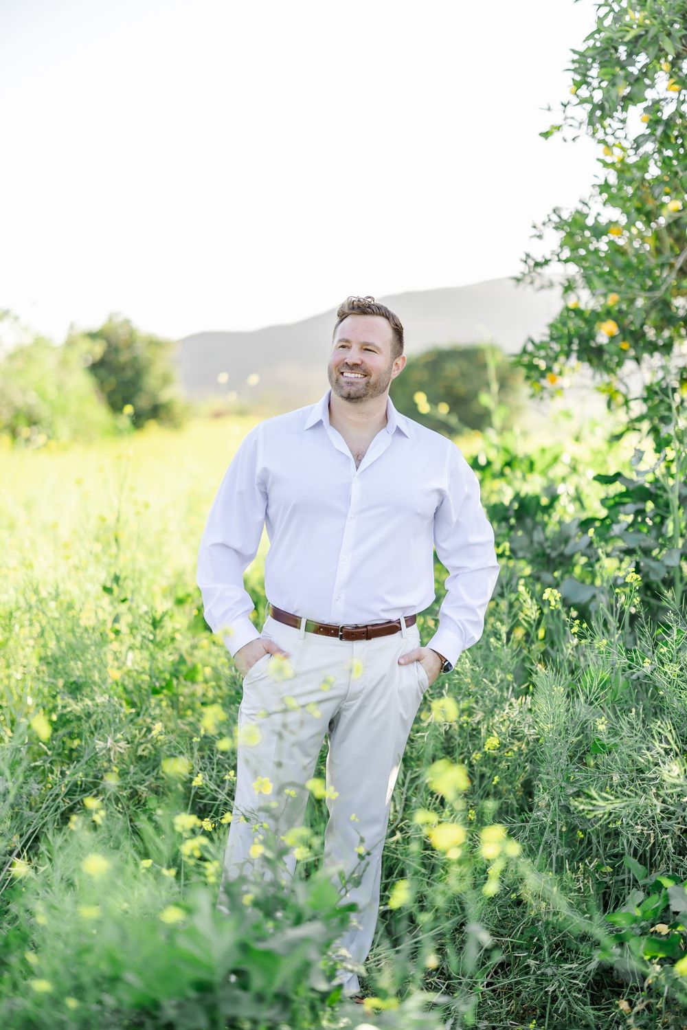 Smiling man in white shirt and khaki pants standing in a lush green meadow with yellow wildflowers and mountains.