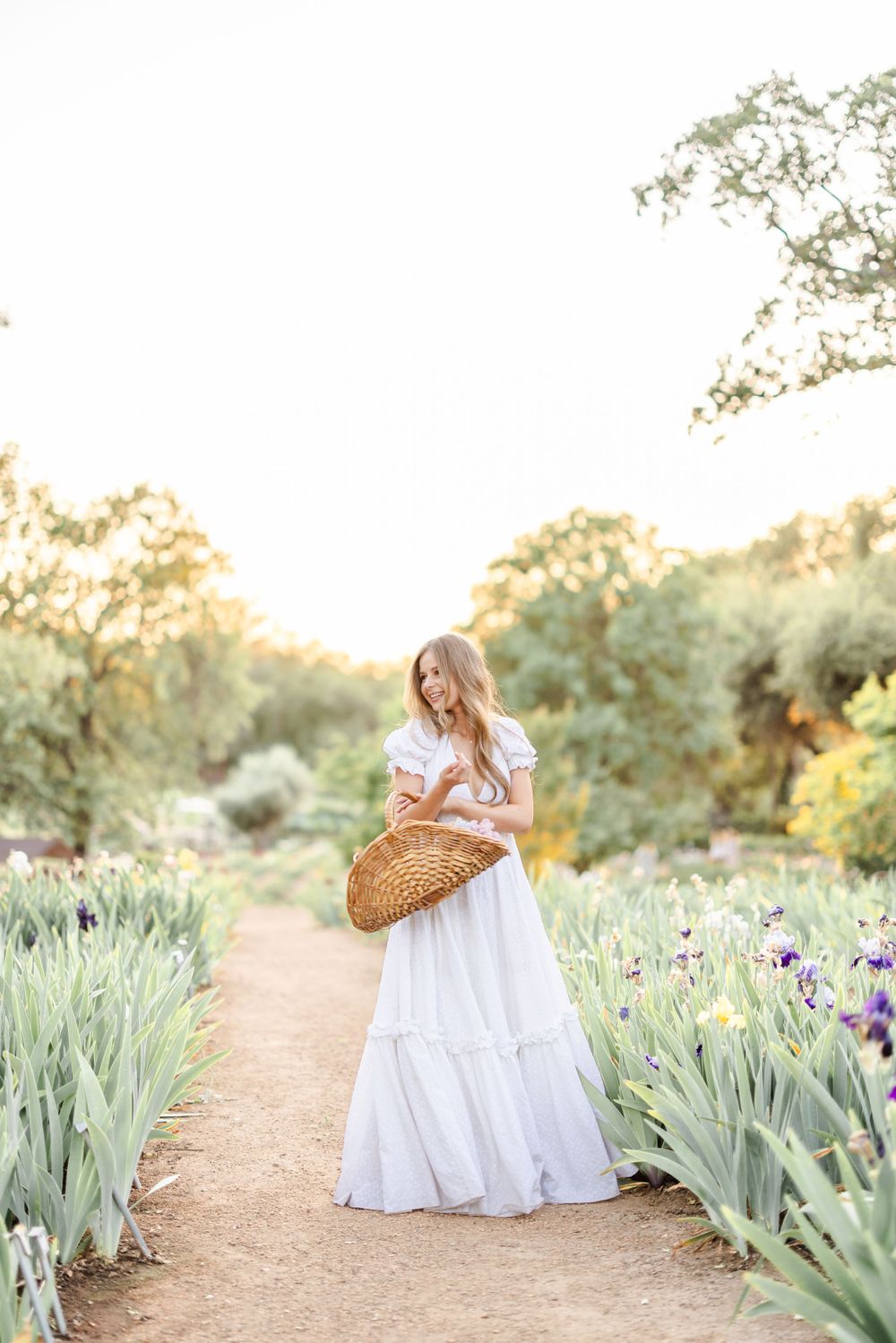 A white-dressed figure walks along a garden path lined with iris flowers carrying a woven basket.