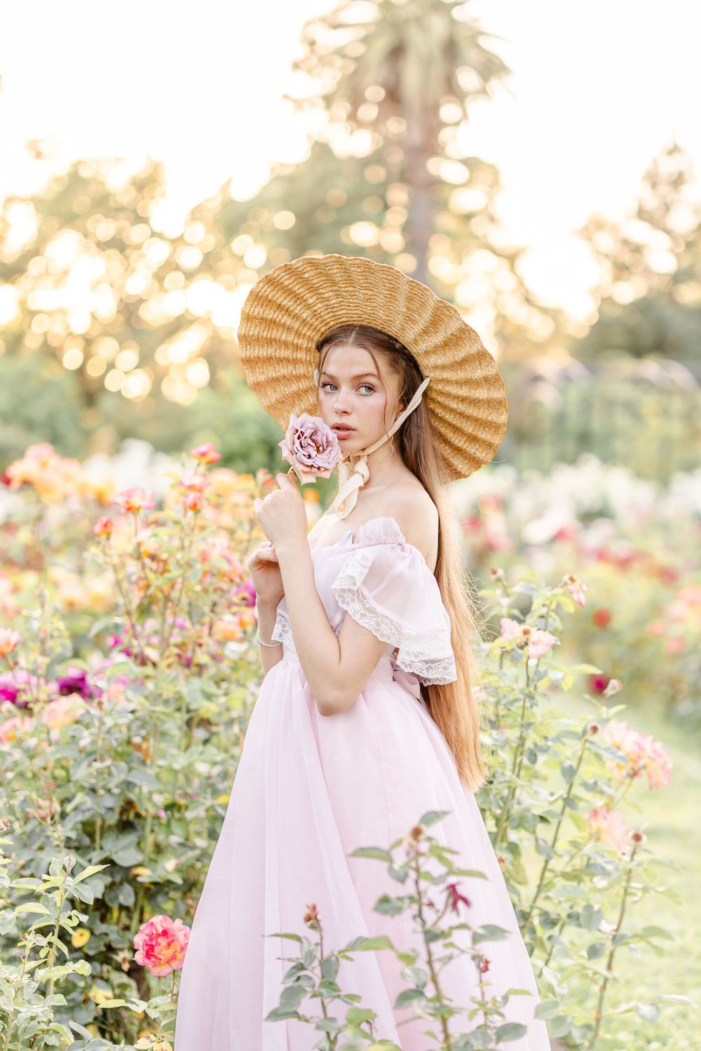 A romantic portrait in a rose garden with soft lighting and a figure wearing a pink dress and woven sun hat.