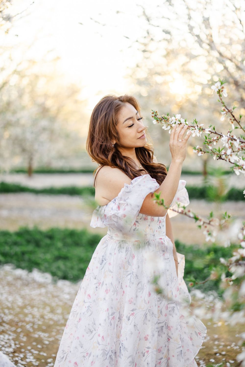 Woman in floral off-shoulder dress smelling cherry blossoms in a sunlit orchard during spring.