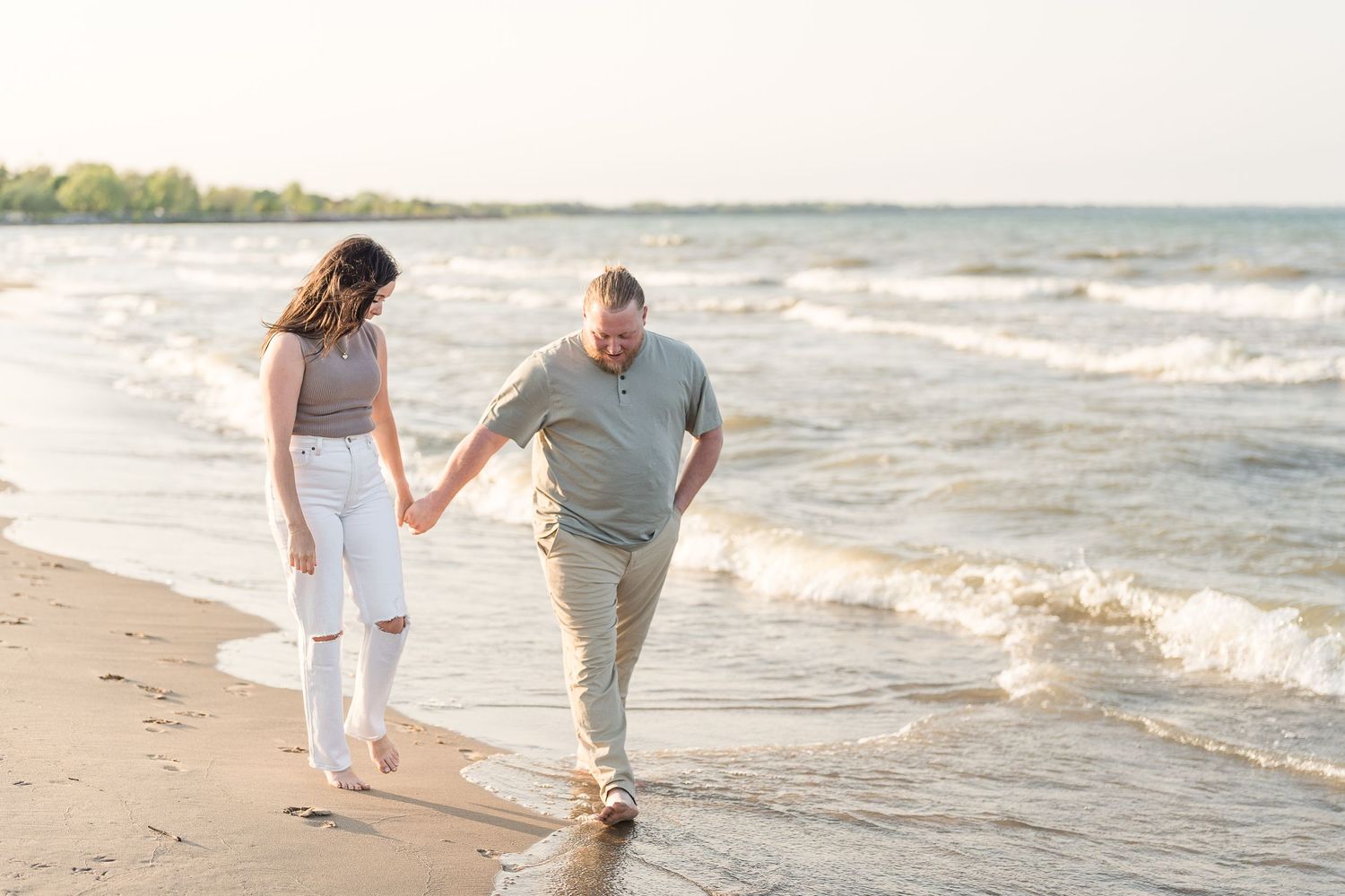 Couple walking hand in hand along a sunny shoreline with gentle ocean waves.