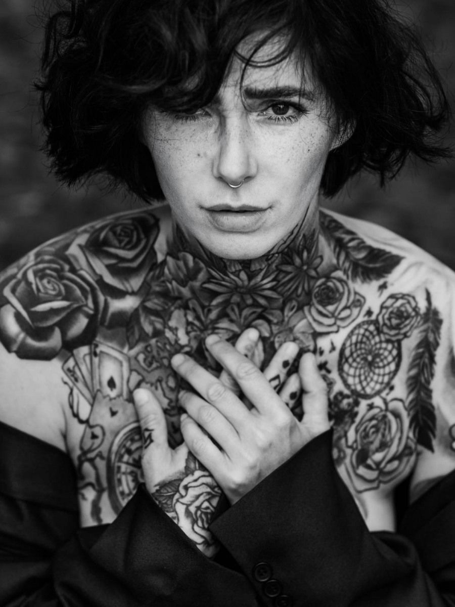 Black and white close-up of a freckled person with extensive chest and neck rose tattoos.