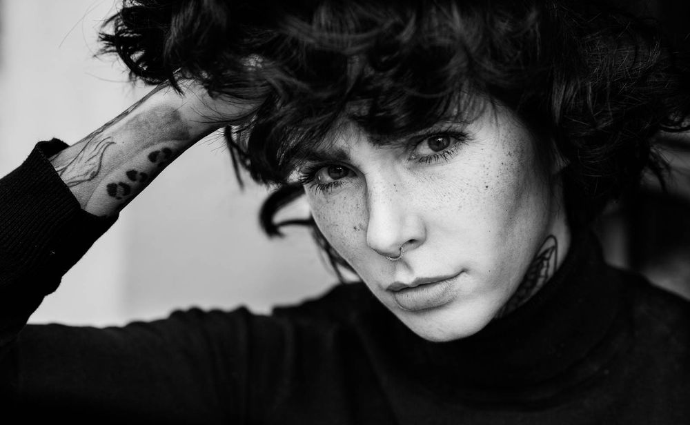 Black and white portrait of a person with short curly hair and tattoos, hand raised to head.