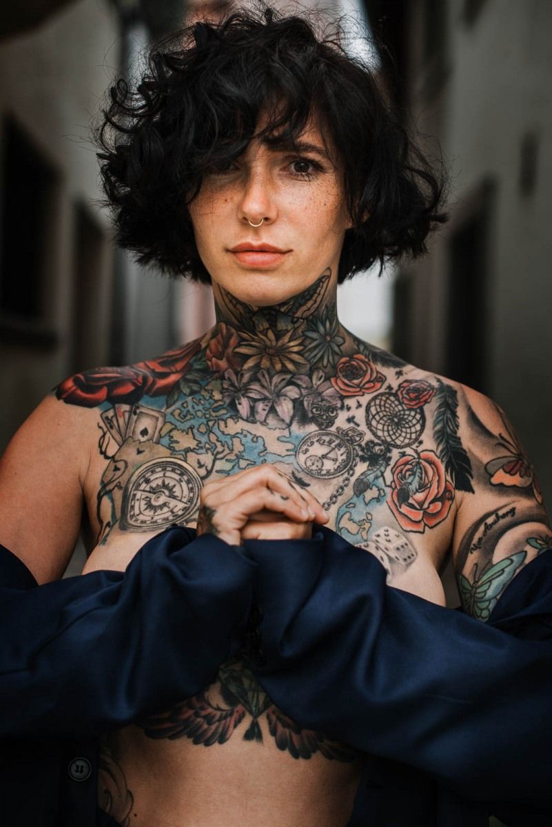 Color portrait of a person with curly hair and extensive colorful chest and neck tattoos.