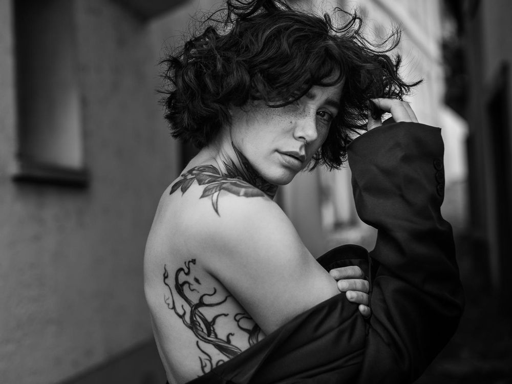 Black and white back portrait of a person with shoulder tattoo draped in dark jacket.