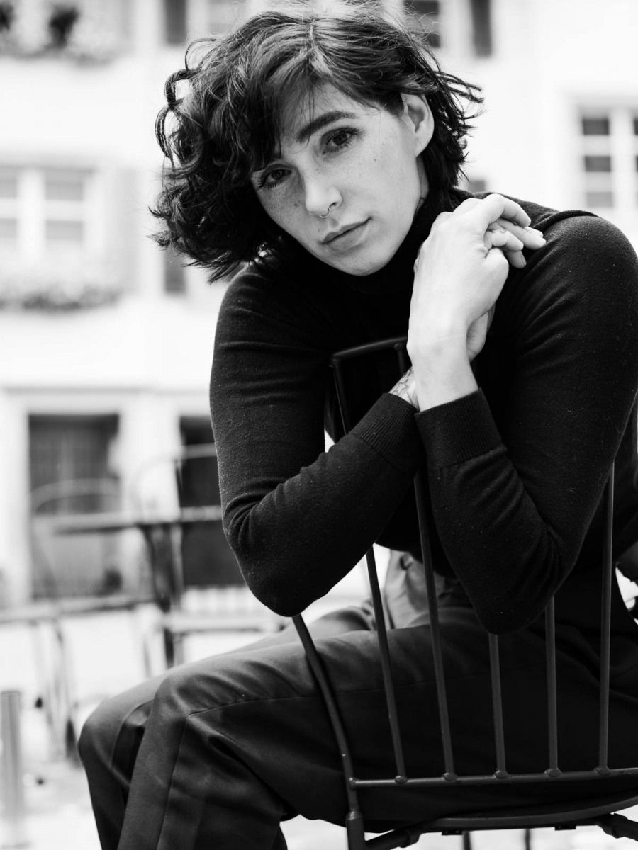 Black and white portrait of a young woman with short curly hair wearing a turtleneck, seated on a chair.