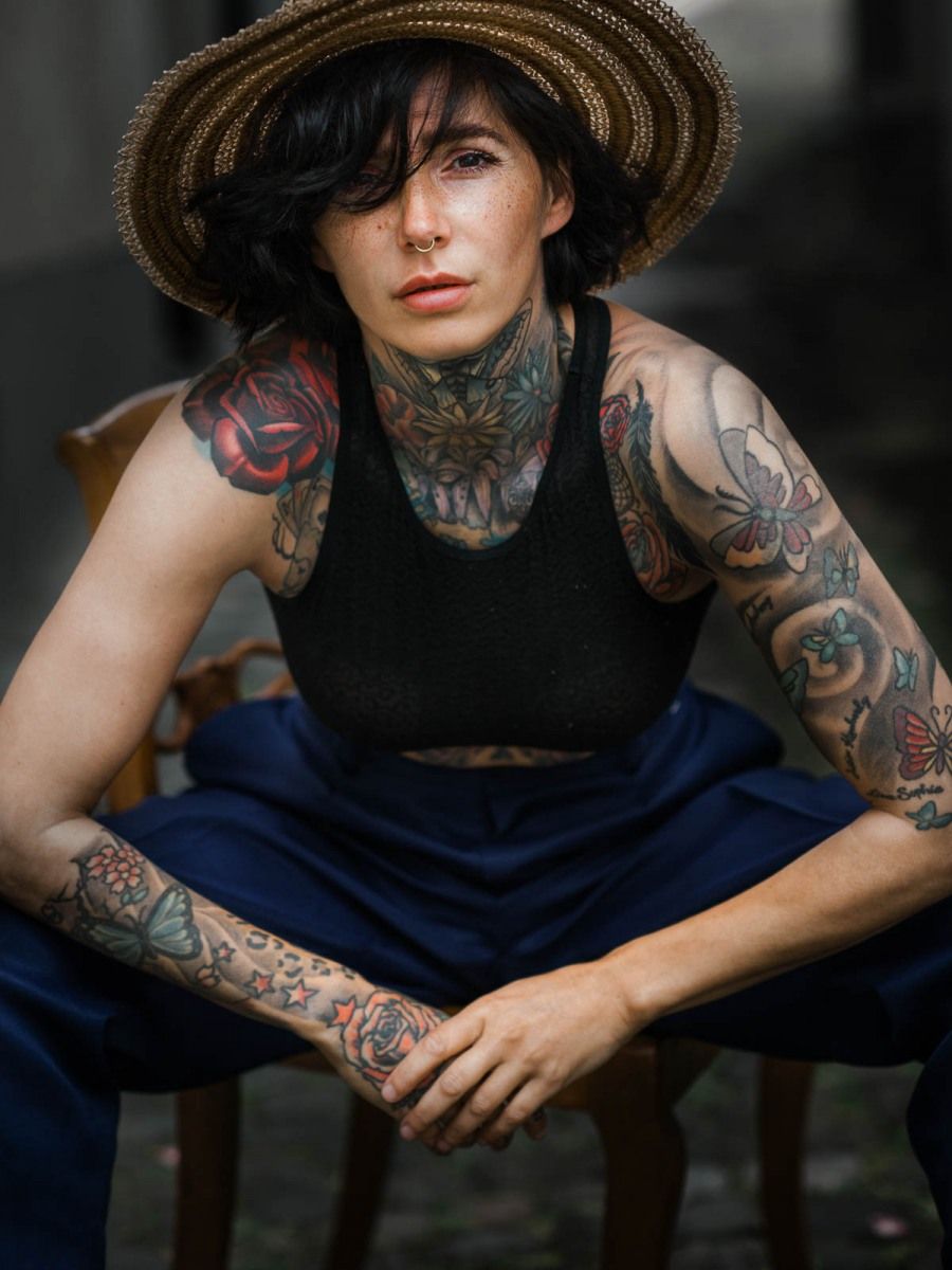 Heavily tattooed woman wearing a straw hat and black crop top, seated confidently in a wooden chair.