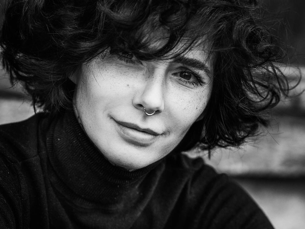 Black and white portrait of a person with curly bob hair and nose ring, soft smile.