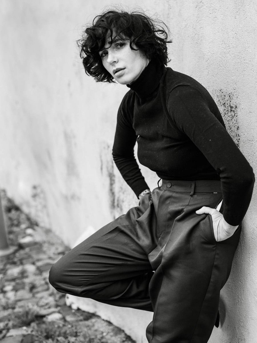 Black and white full body shot of a person in turtleneck and wide-leg pants leaning on wall.