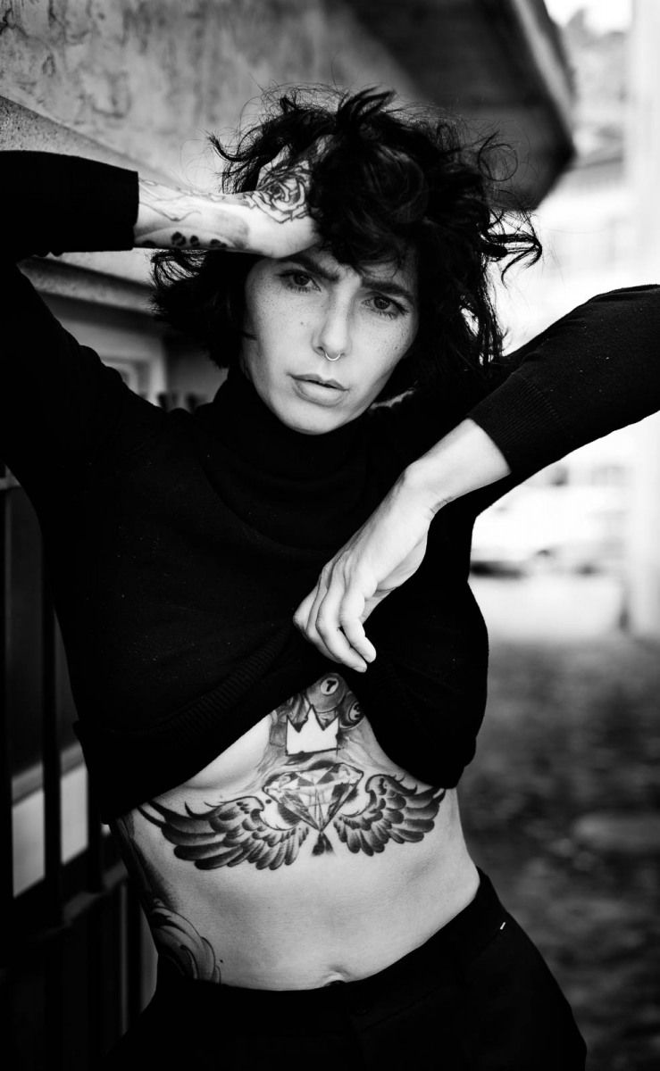 Black and white photo of a tattooed person in black top lifting shirt to reveal stomach tattoo.