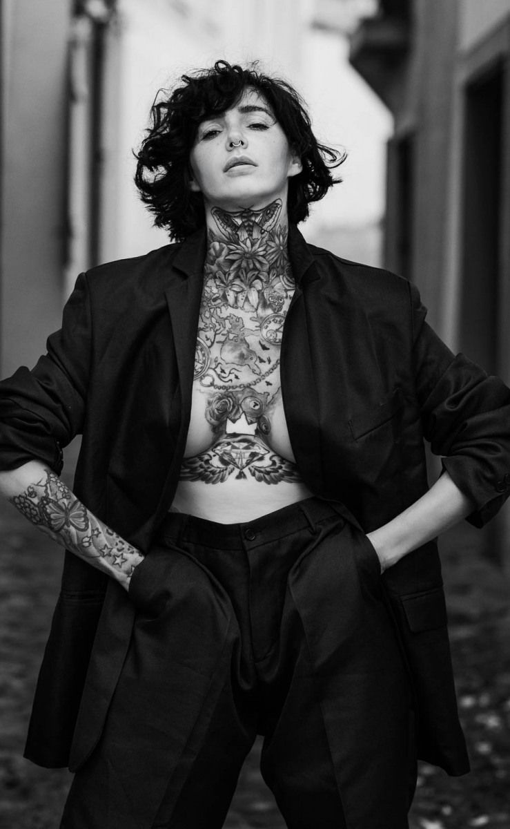 Black and white photo of heavily tattooed person in open blazer posing on cobblestone street.