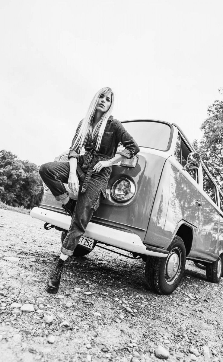 Black and white photo of blonde woman posing on vintage small car in outdoor setting.