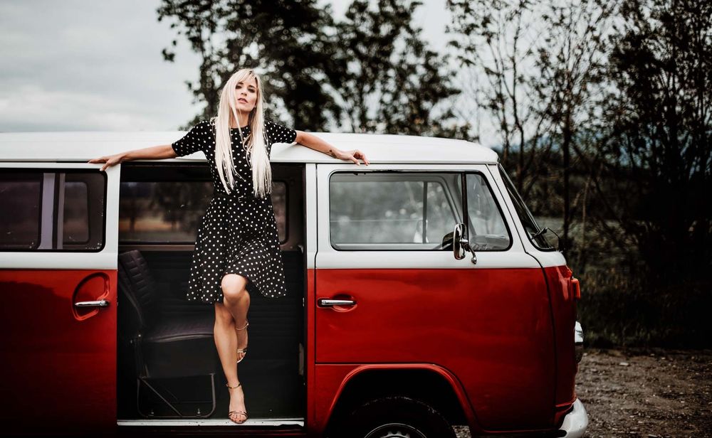 Woman in polka dot dress leaning against red and white vintage Volkswagen van outdoors.