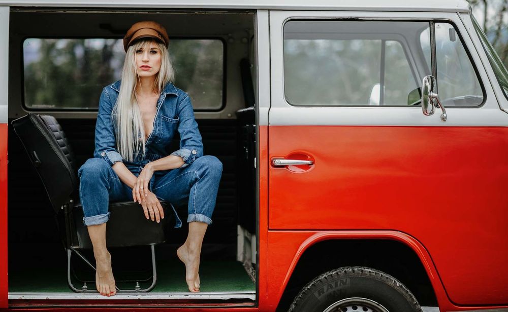 Blonde woman in denim jumpsuit sits in red vintage VW van doorway.