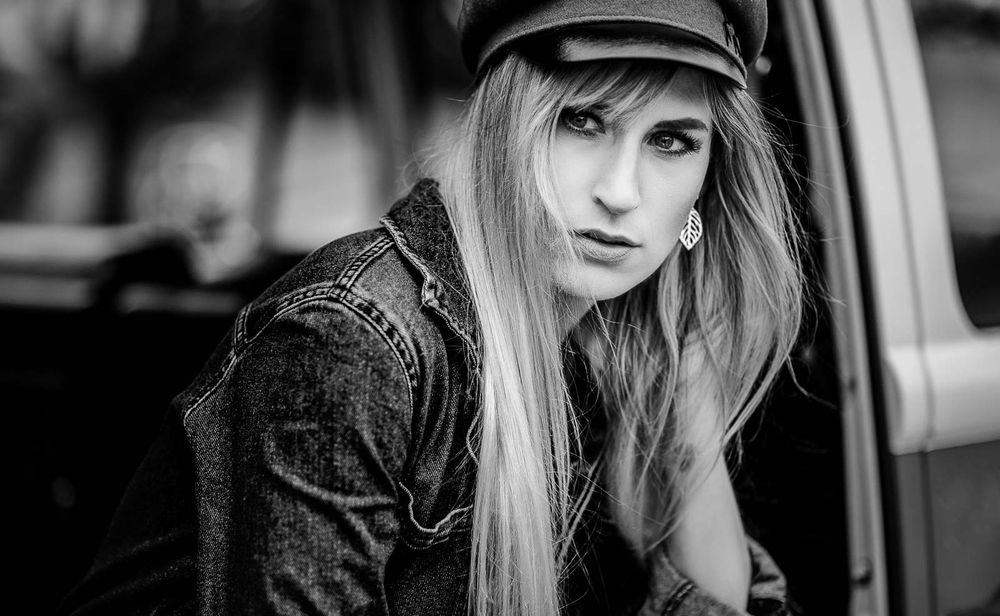 Close-up black and white portrait of blonde woman in newsboy cap near car.