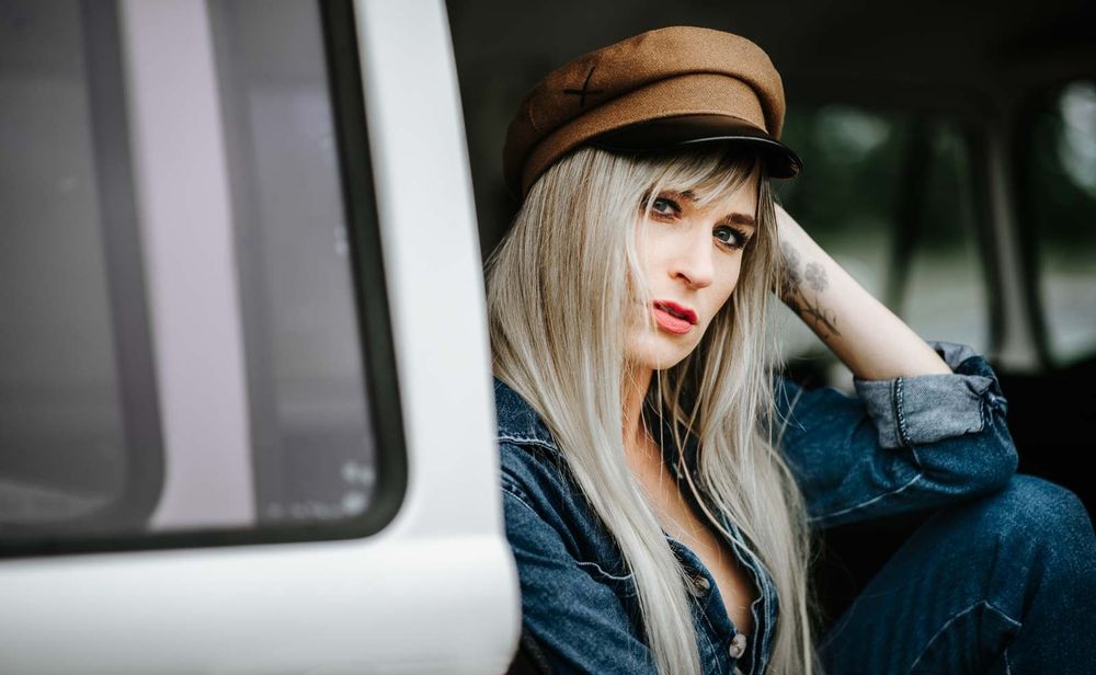 Blonde woman in brown cap and denim jacket leans against white vintage van.