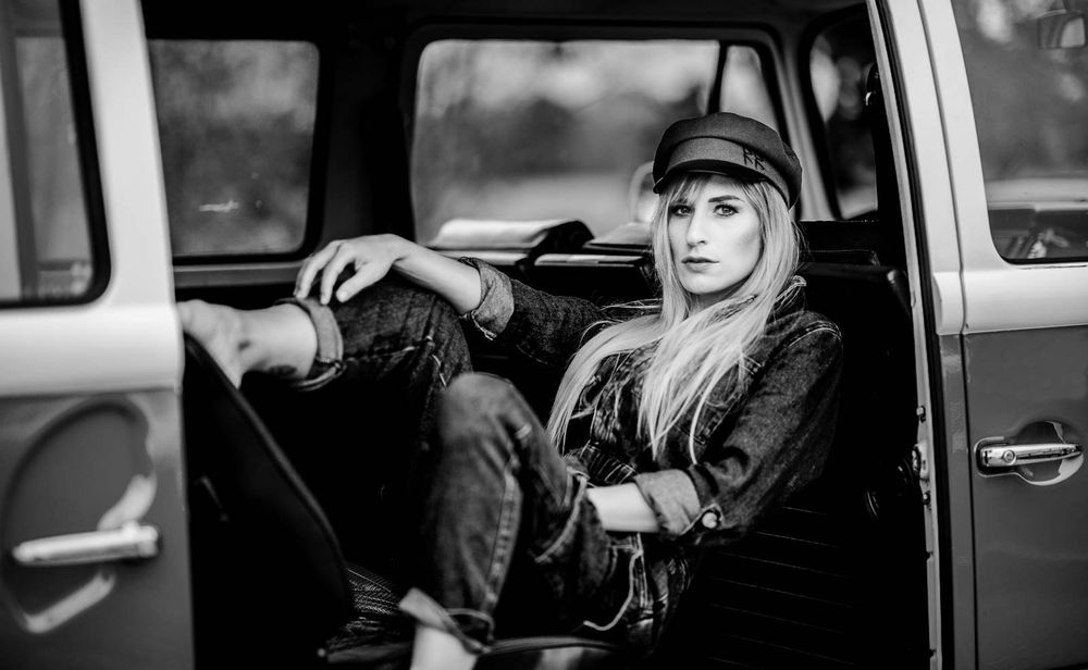 Woman in black outfit and hat sits cross-armed in vintage van, black and white.