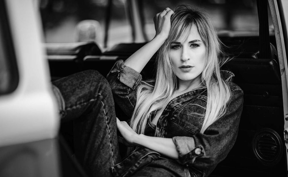 Blonde woman in denim sits inside vintage vehicle, black and white portrait.