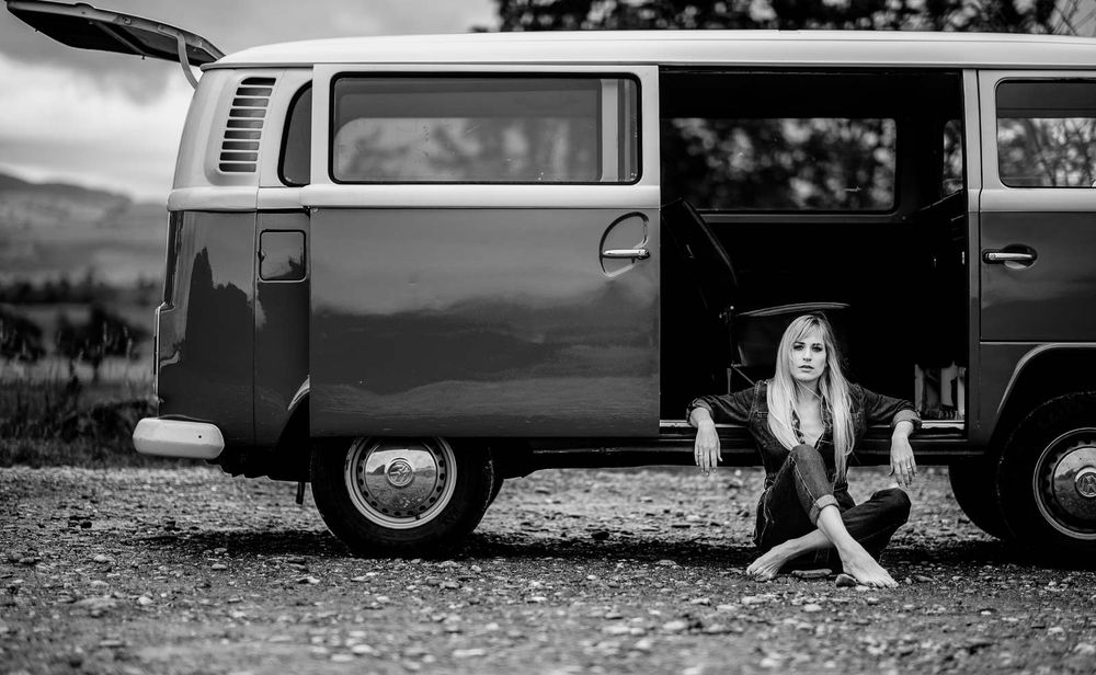 Young blonde woman sits beside classic VW camper van on gravel, black and white.