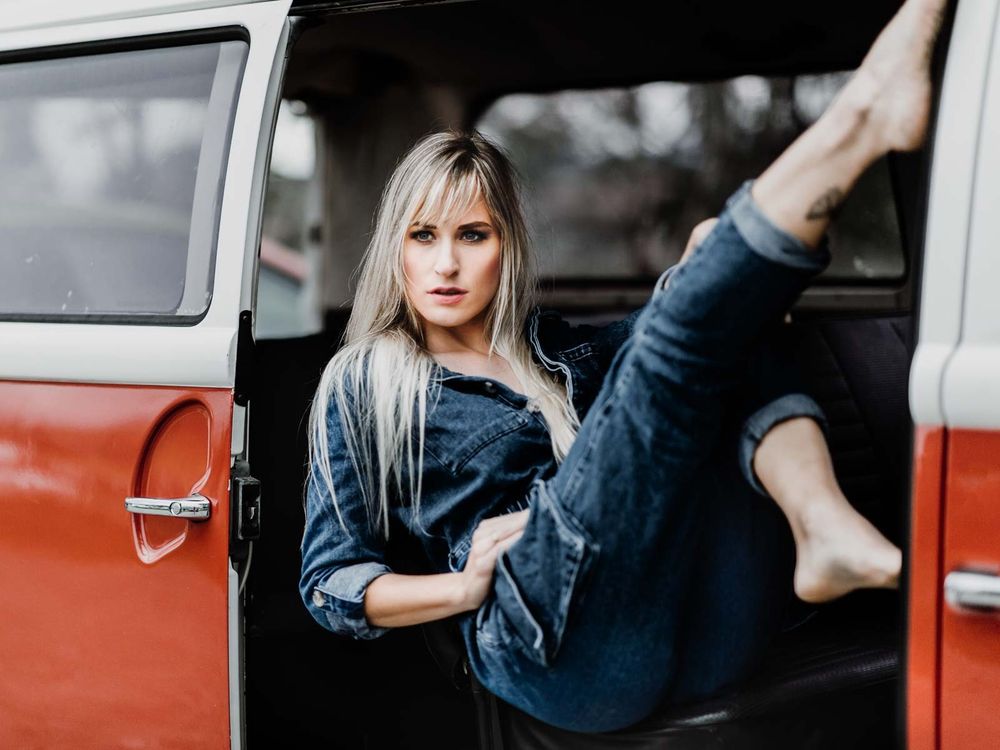 Barefoot blonde woman in denim poses with leg raised in red VW van doorway.