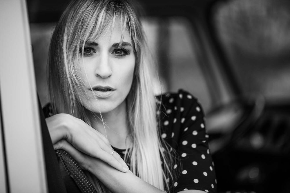 Close-up black and white portrait of blonde woman in polka dot top near car window.