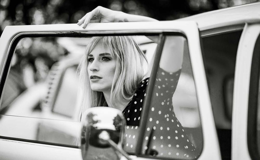 Blonde woman in polka dot dress gazing from vintage car window in black and white photo.