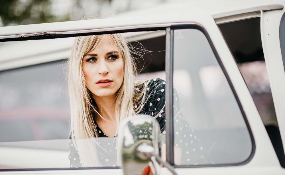 Young blonde woman looking through old white vehicle window wearing black polka dot dress.