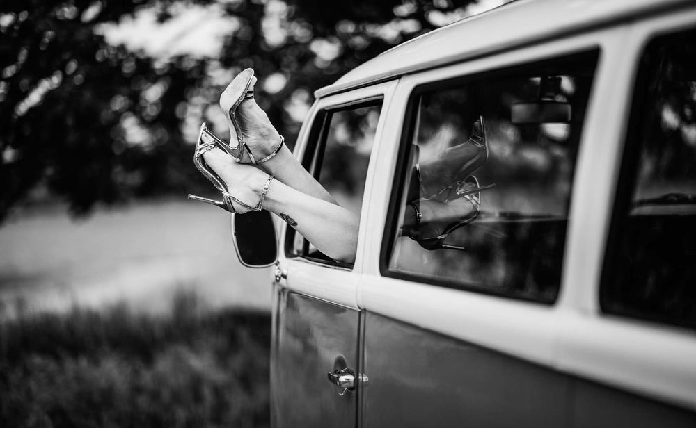 Black and white photo of high heels dangling from vintage van window outdoors.