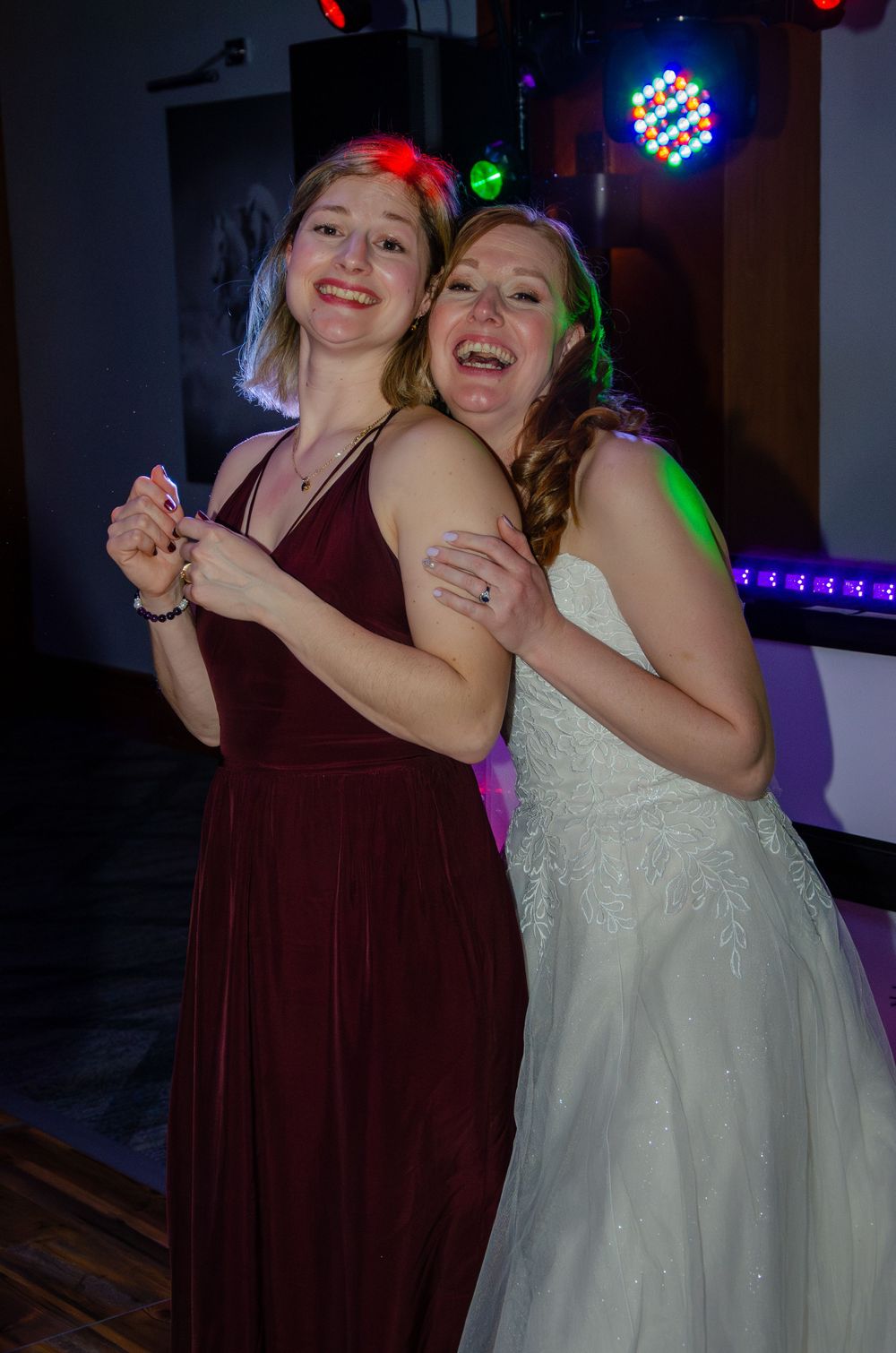 Two wedding guests embrace and laugh on the dance floor under colorful party lights.