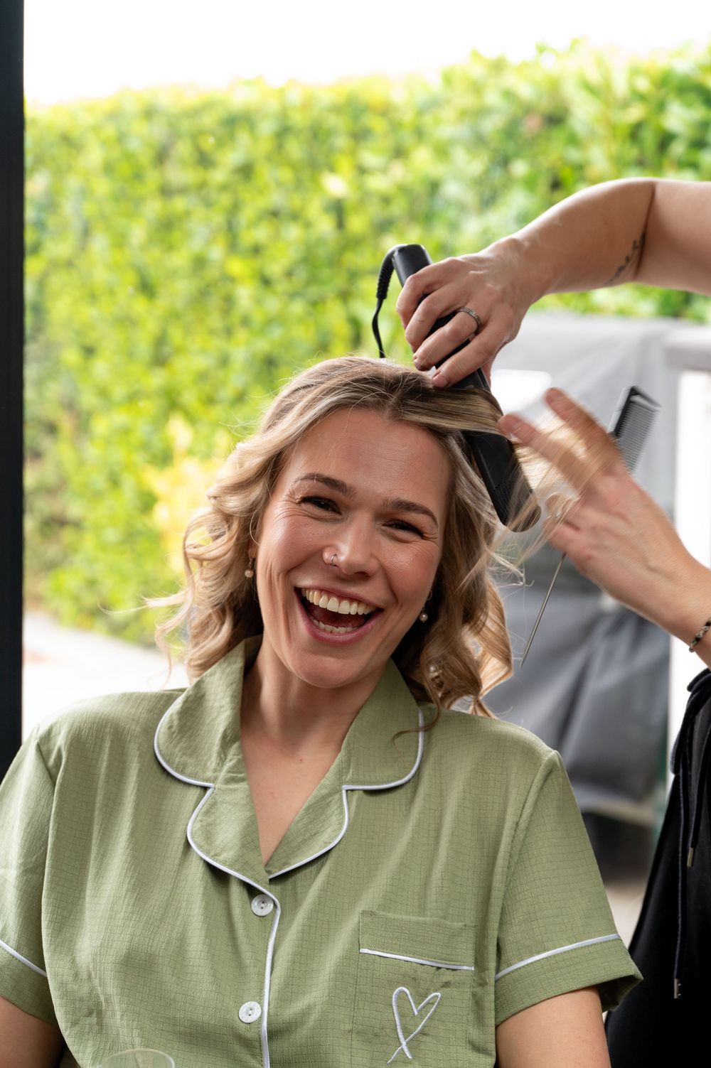 Someone wearing a sage green pajama-style top with white piping laughs joyfully while getting their hair styled.