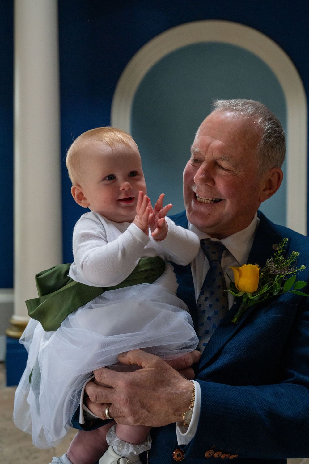 A baby in a white dress smiles while being held by an older person wearing a blue suit with yellow boutonniere at formal event.
