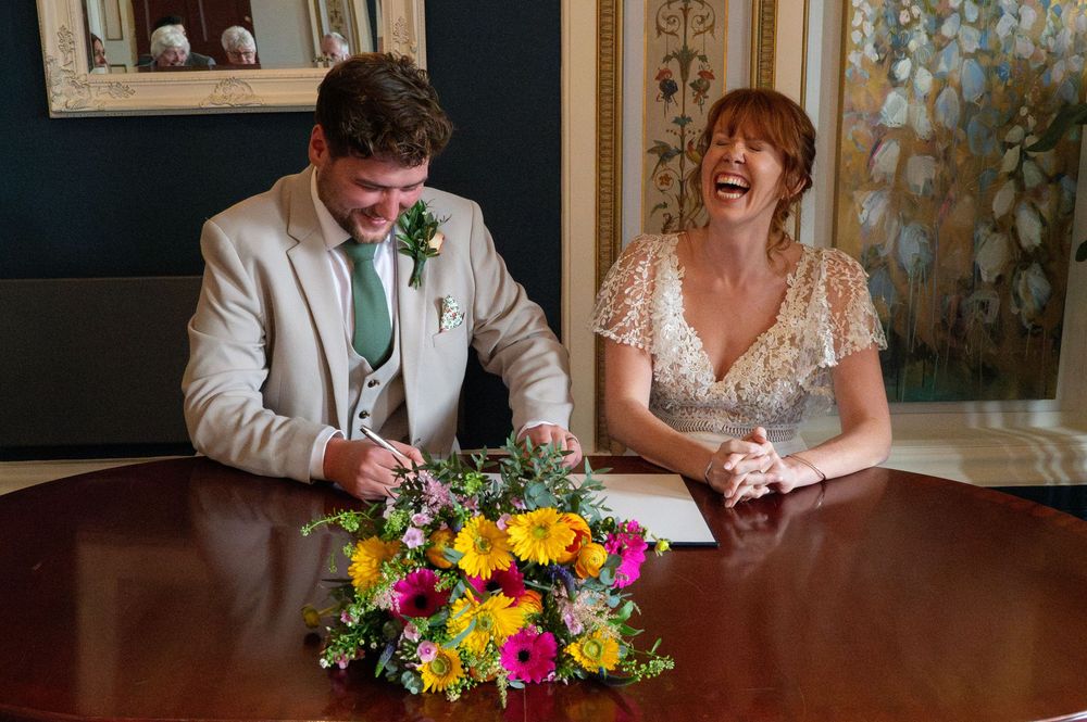 A couple signs marriage paperwork at a wooden table with a vibrant wildflower bouquet between them.