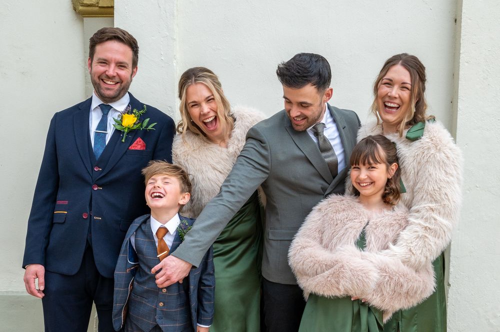 A joyful group laughs together while posing against a white wall at a formal event.
