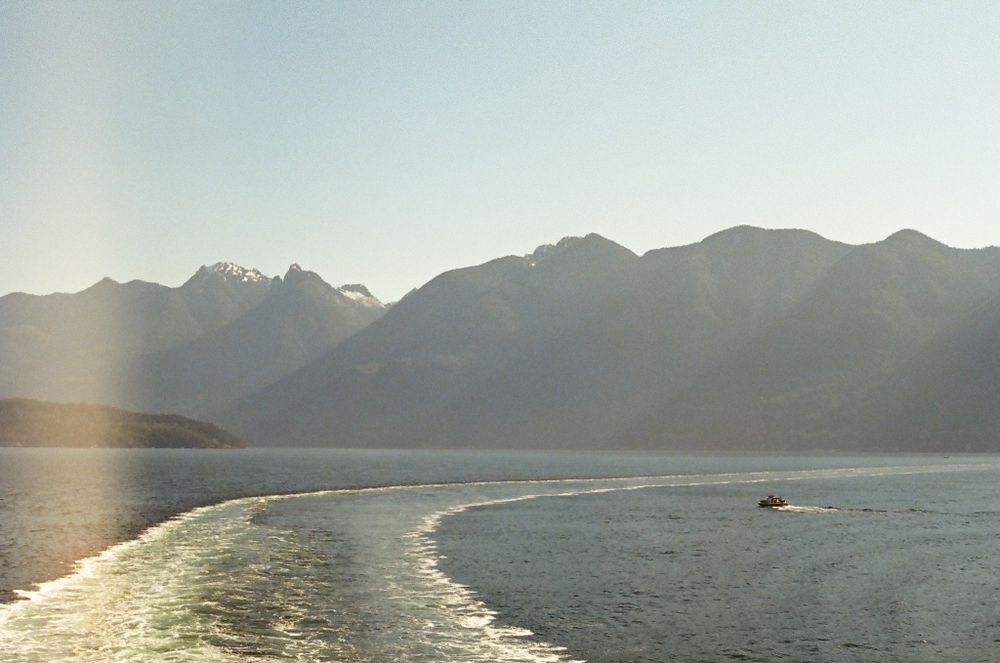 Misty mountain silhouettes across water with ferry wake visible in golden sunlight.