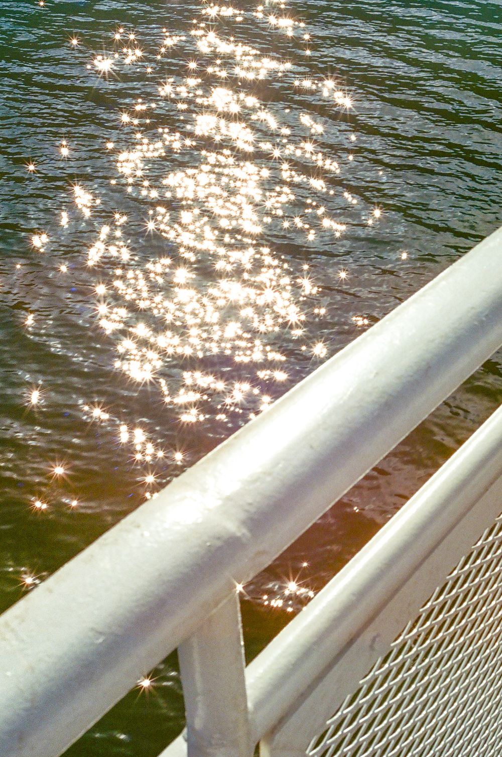 Sunlight sparkles on rippling water surface viewed through a white boat railing.