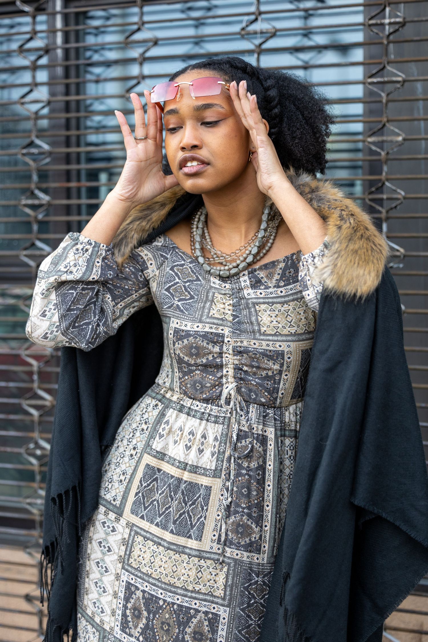Fashionable Black woman posing with pink sunglasses wearing patterned maxi dress and fur-trimmed wrap outdoors.
