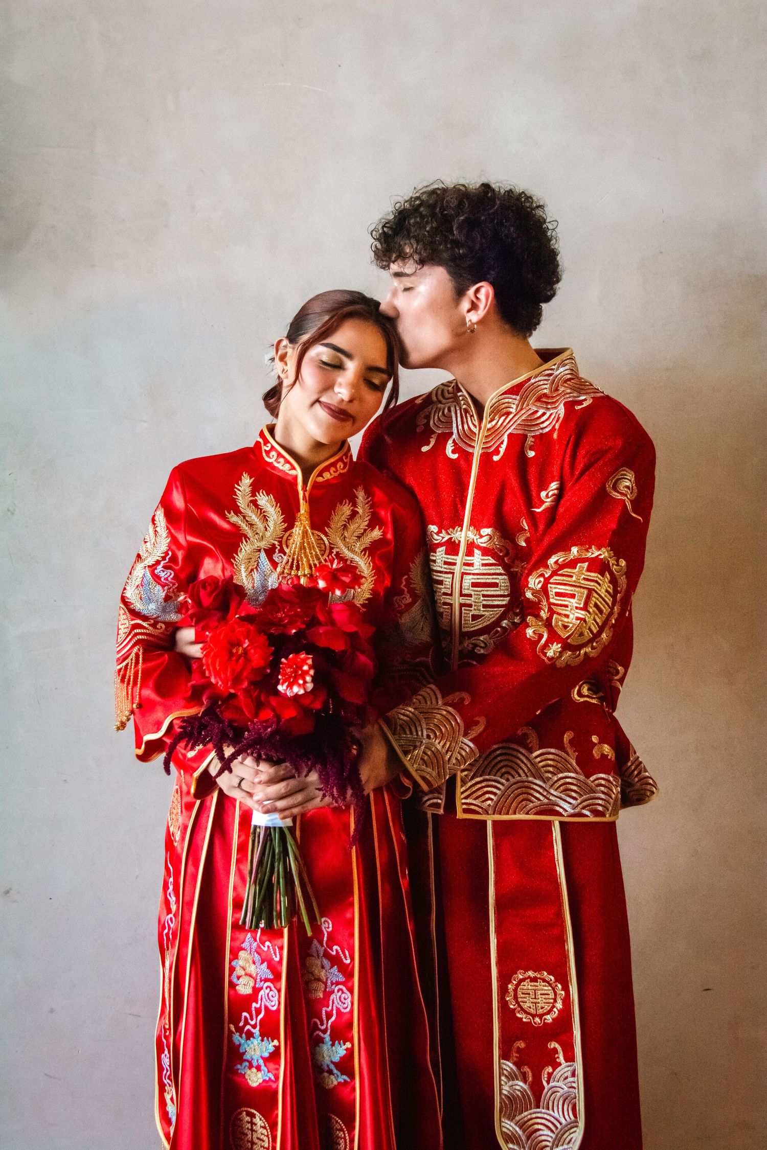 Couple in traditional red Chinese wedding attire embracing while bride holds a red floral bouquet.