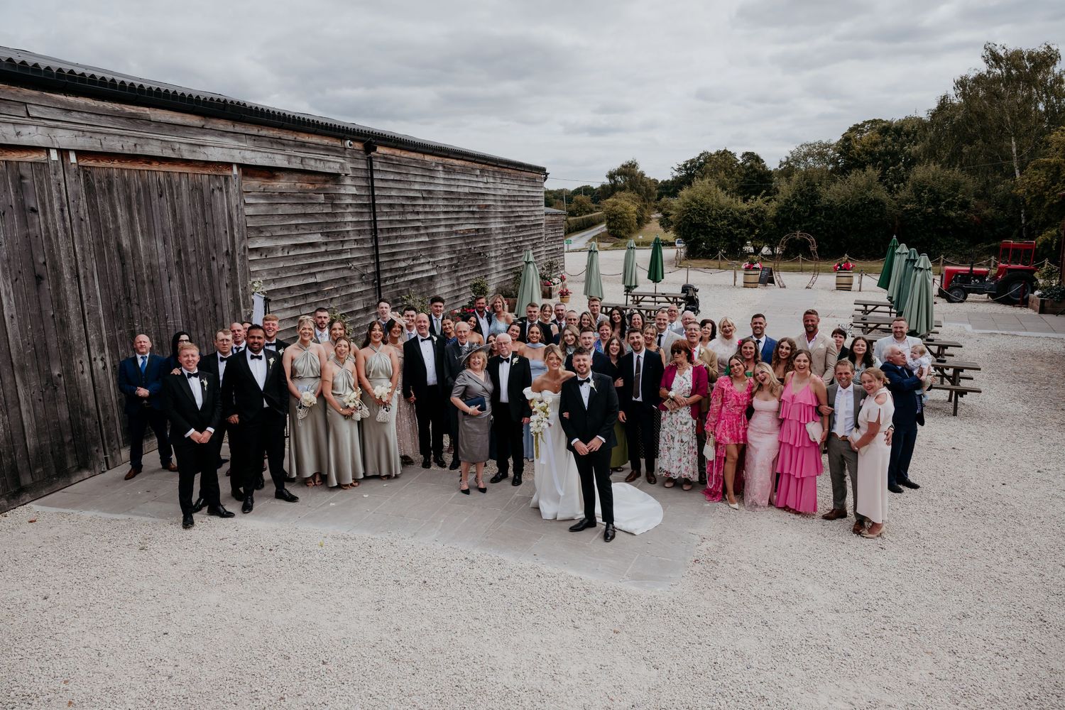 Panoramic wedding party group photo taken outside a rustic venue.