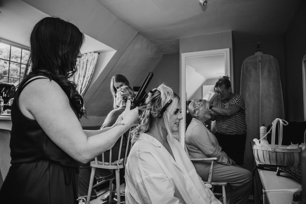 A black and white photograph of bridal preparations, showing hair and makeup styling in a well-lit room. Houchins