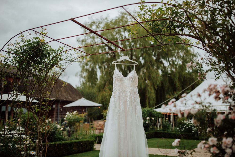 White wedding dress hangs from arched garden trellis with lush greenery and outdoor venue setting in background.