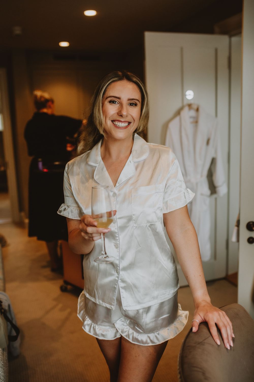 A person in white silk pajama shorts set holds a champagne glass while getting ready in a warmly lit room.