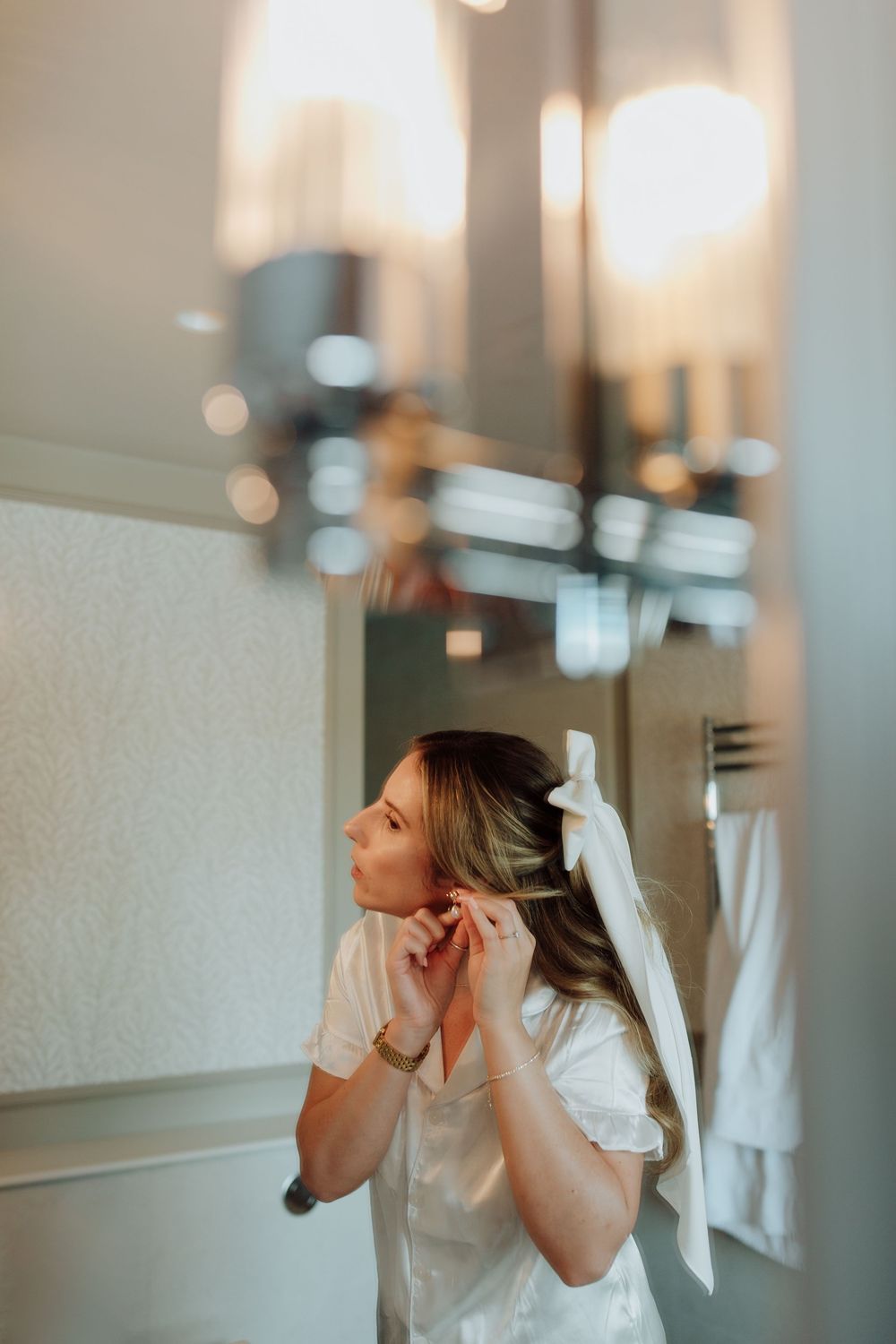 Person in white clothing looks at reflection in bathroom mirror with soft bokeh lighting effect in background.