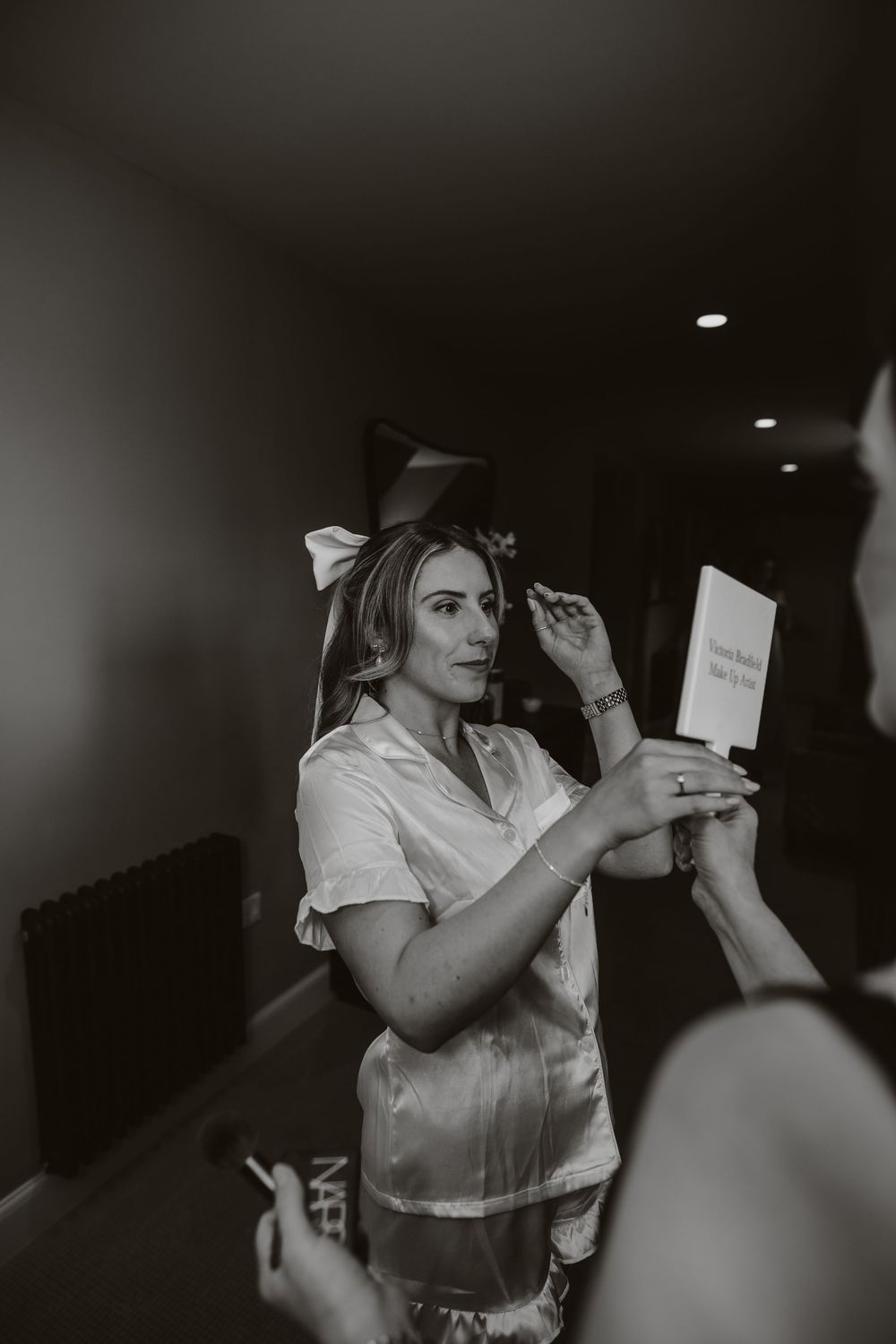 Black and white photo of a woman in a light-colored dress shirt and bow getting ready in a dimly lit room.