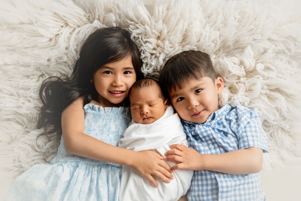 Two older siblings in blue outfits lovingly hold a swaddled newborn baby on a fluffy white rug.