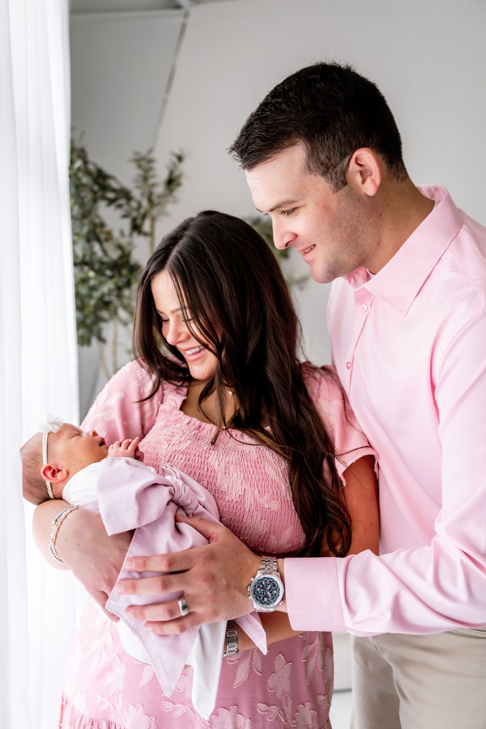 A couple in pink outfits smiling at their newborn baby wrapped in a lavender blanket during a newborn photo session.