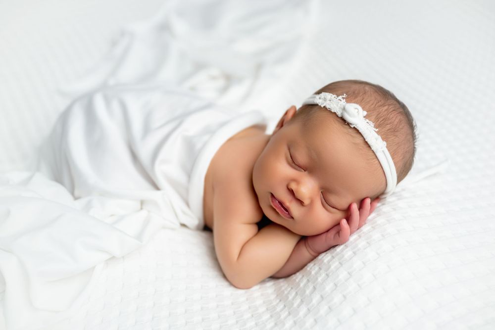 Peaceful newborn baby sleeping wrapped in white blanket with floral headband on white background.