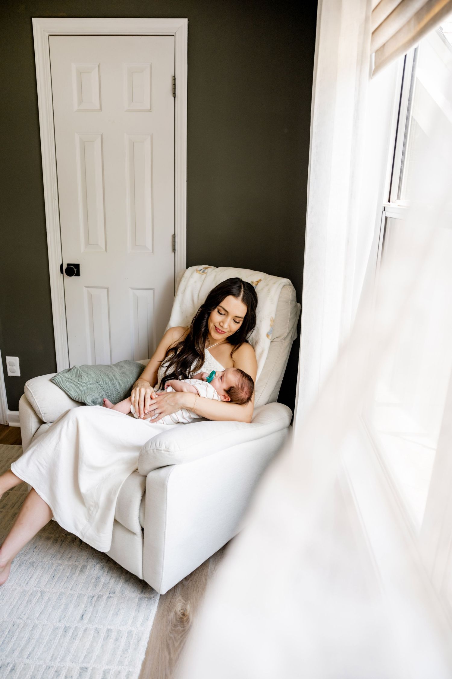 A mother sits in a white recliner chair holding her newborn baby near a bright window in a cozy room.