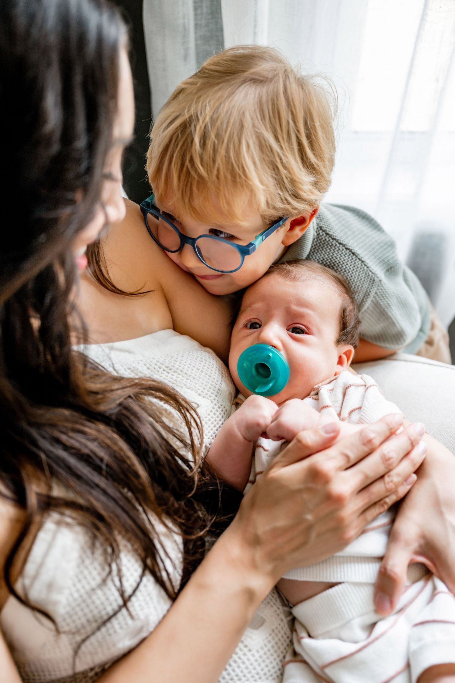 A mother holds a newborn with a pacifier while an older child with glasses leans in to kiss the baby.