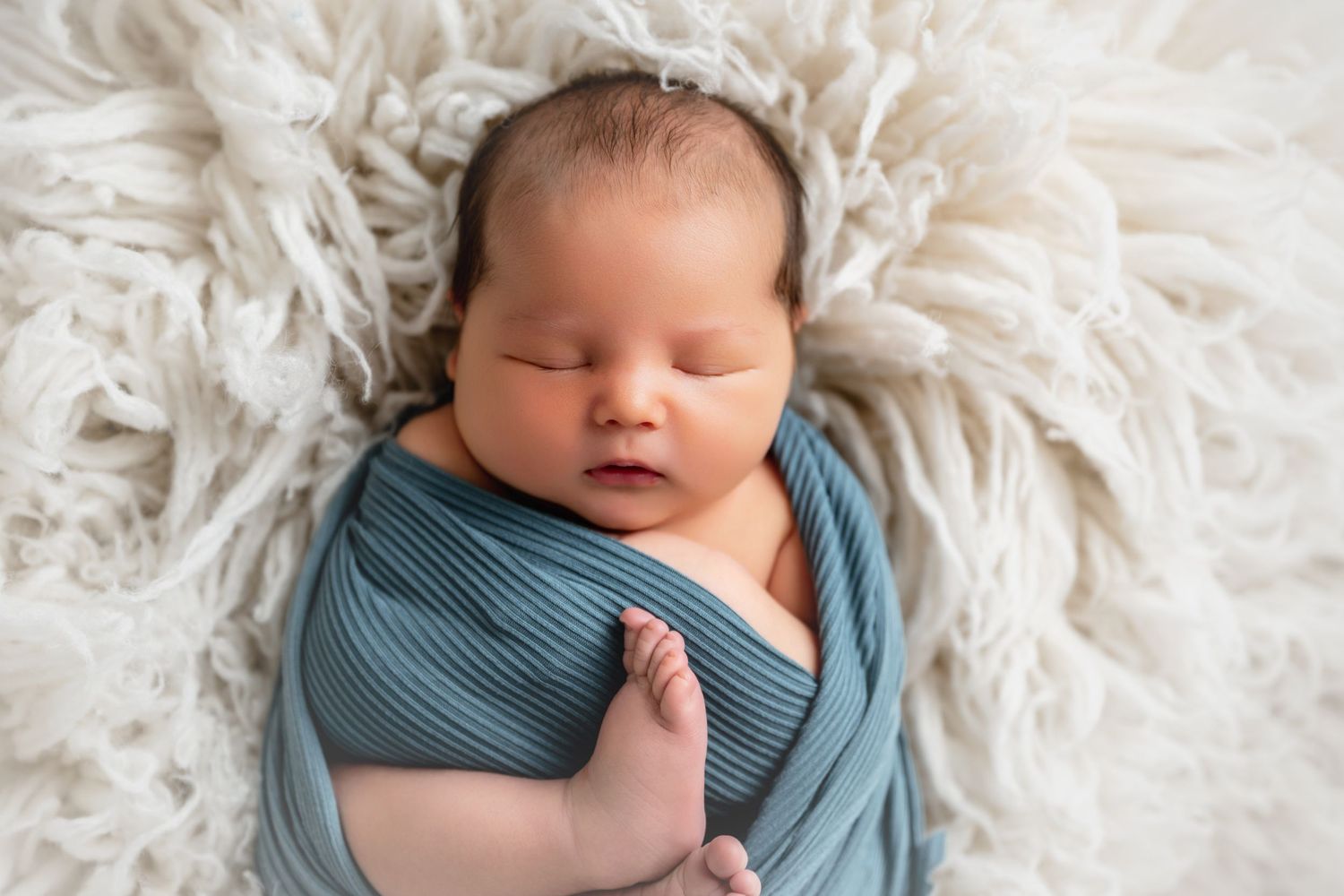 Peaceful newborn baby swaddled in a teal wrap, sleeping on a soft cream fluffy blanket.